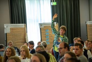 Ein junger Mann meldet sich beim Bürgerinnenrat. Im Hintergrund sieht man beschriftete Flipcharts