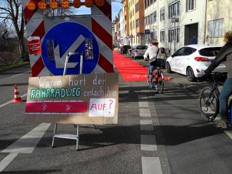 Menschen fahren Fahrrad auf Rotem Teppich, den den Radschutzstreifen verlängert. Daneben ein Schild mit der Aufschrift "Warum hört der Fahrradweg einfach hier auf?