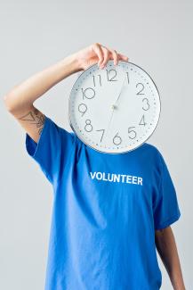 Mensch mit einem blauen T-Shirt und der Aufschrift "volunteer" hält sich eine Wanduhr vor's Gesicht.