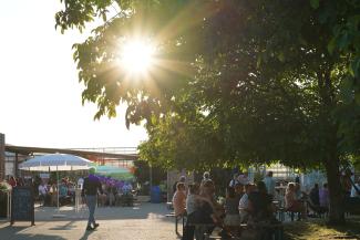 Hof der Gärtnerei Piluweri mit Menschen an Tischen; Abendsonne im Hintergrund